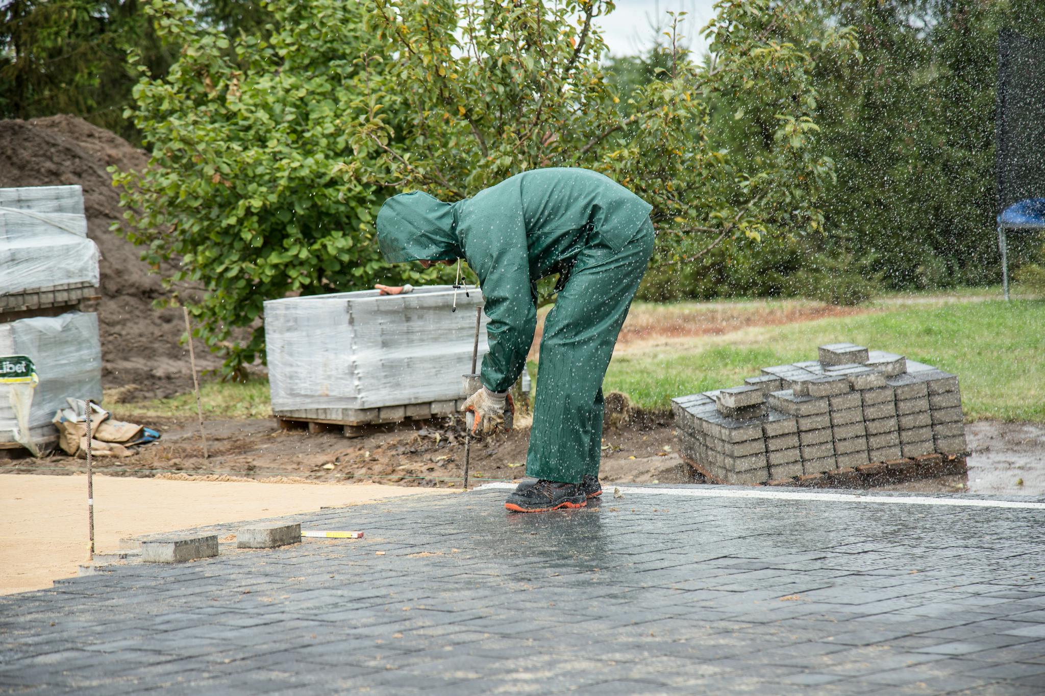 A worker in protective gear lays paving stones on a wet day.